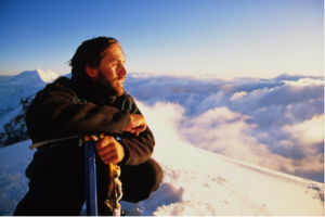 A photo of Erik Weihenmeyer, looking off into the distance while sitting on a snowy mountain peak, surrounded by mist, clouds and a blue sky.