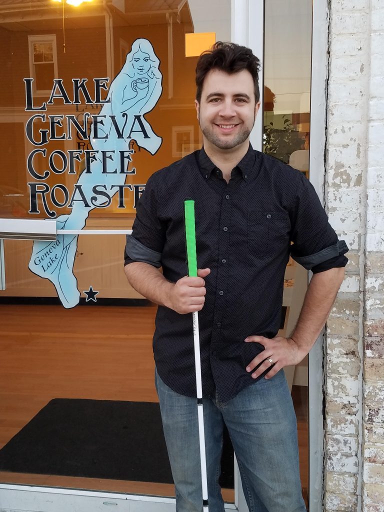 Jeremiah Fox, smiling and holding a mobility cane with a green handle, stands at the Lake Geneva Coffee Roastery storefront