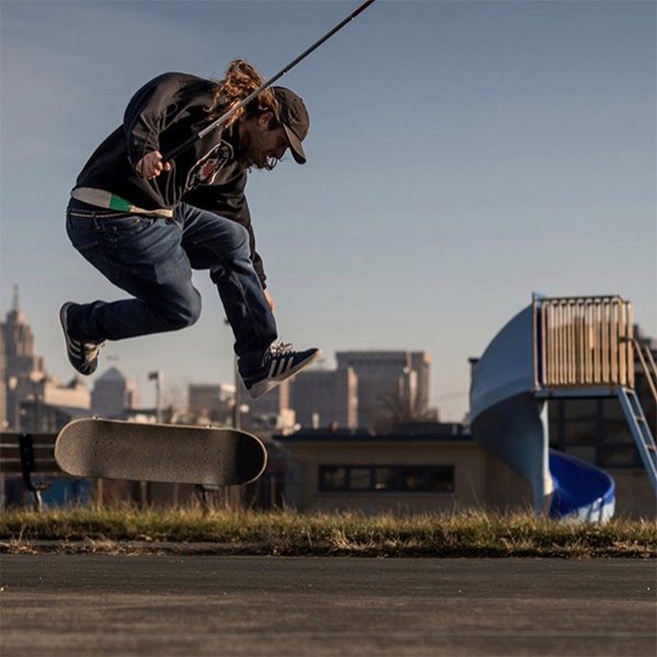 Dan Mancina is skateboarding in an urban park - holding his white cane in his right hand while jumping high into the air as his skateboard spins below him.