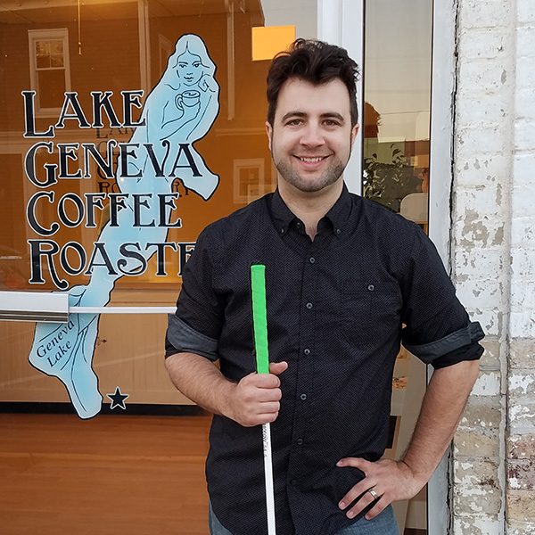 Jeremiah Fox, smiling and holding a mobility cane with a green handle, stands at the Lake Geneva Coffee Roastery storefront