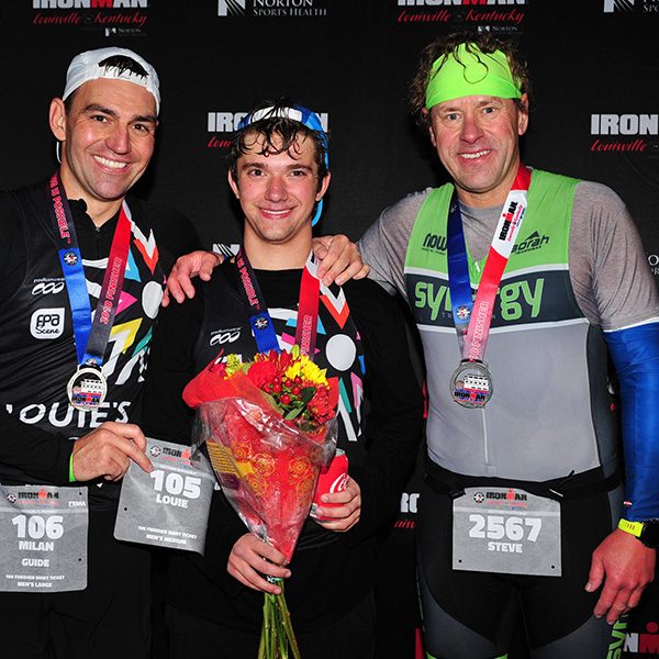 Louie McGee (center) smiling with his IRONMAN medal and a bouquet of flowers. On his left is his race guide, Milan Tomaska, and on his right is family friend Steve Roeske - both of whom helped Louie through the last 12+ miles when he had no vision at all.