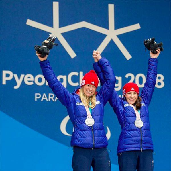 Jen and Menna at the PyeongChang 2018 Winter Paralympics dressed in blue coats and red hats, wearing their silver medals while holding hands and raising their arms in victory.