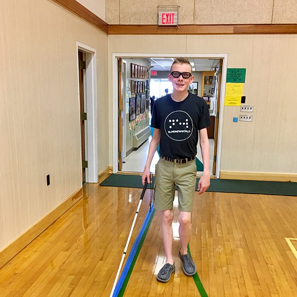 Noah Carver, wearing a BlindNewWorld t-shirt, practicing for his 8th grade celebration procession and recession using a rope taped down with painters tape as a guide to independently march into the gymnasium, through the music room and onto a stage.