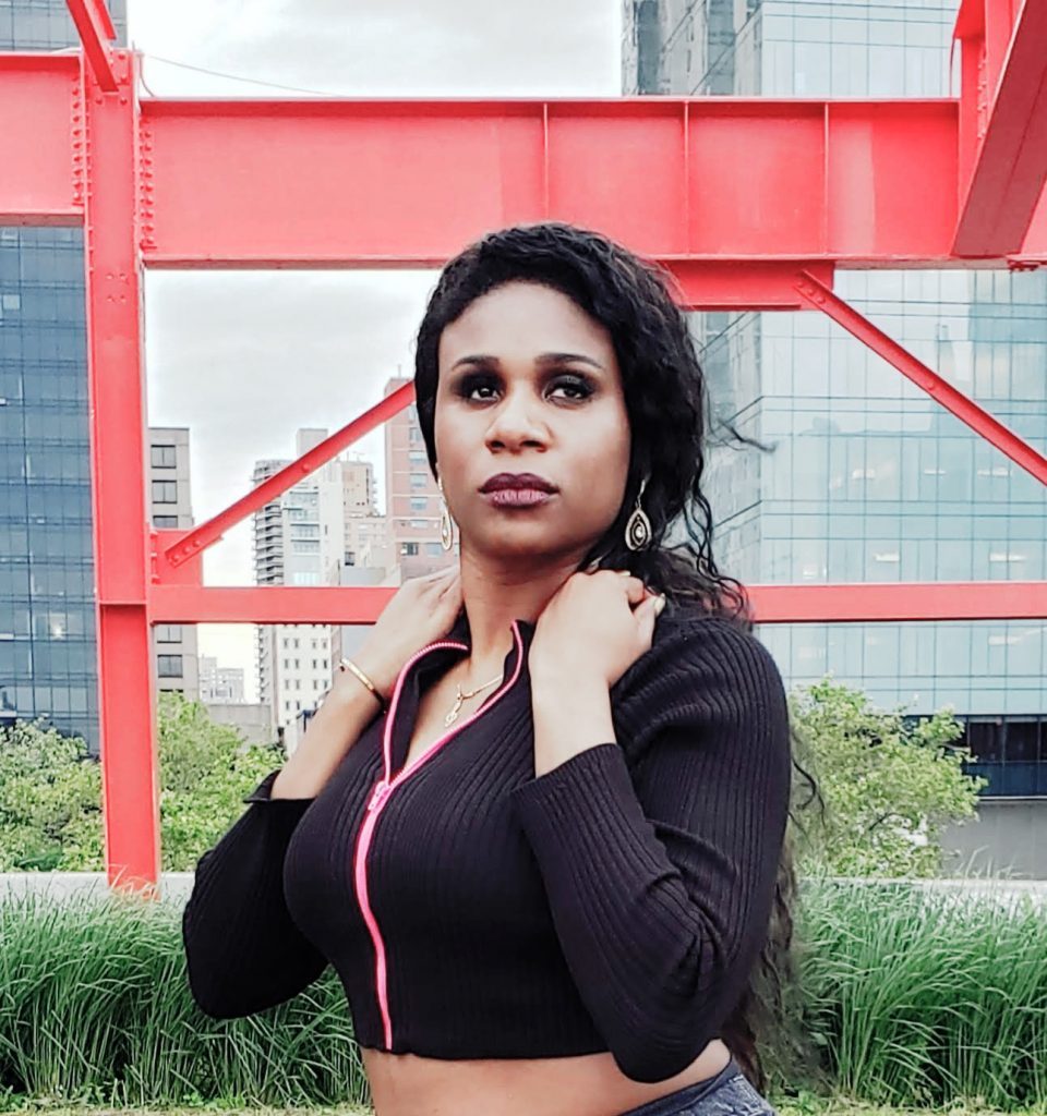 Lachi, wearing dangly earrings and a cropped black sweater with a hot-pink zipper, stands in front of red metal scaffolding and greenery in New York City. She's looking confidently at the camera.