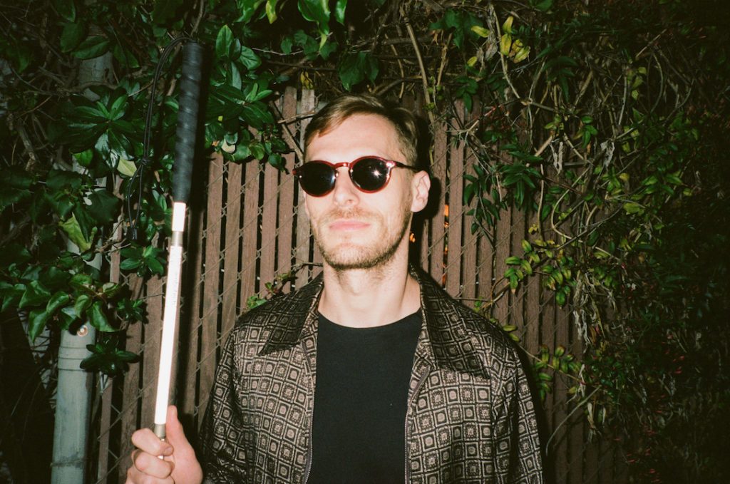 Casey Harris stands in front of a wooden fence surrounded by greenery. He's wearing red, dark glasses and holding his mobility cane in his hand while looking confidently at the camera.