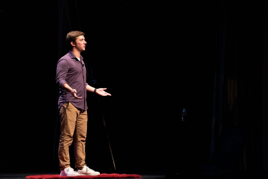 Louie, wearing a purple button-down and khakis, stands in his “red dot” on the TEDx stage