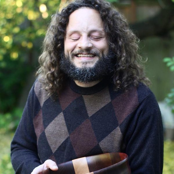 John, sporting a dark beard and long dark curly hair is wearing a dark sweater and holding a large wood bowl. The bowl is dark with a light colored stripe through the middle. Bowl is made of Black Walnut, Padauk and Ash woods.