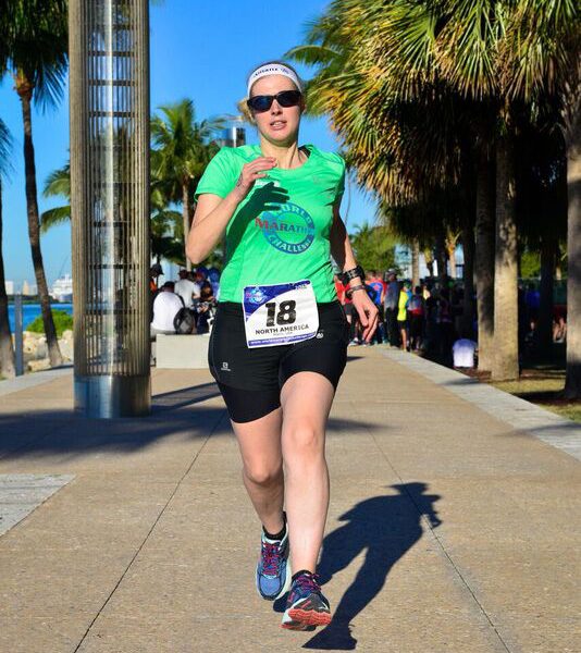 Sinead, wearing a bright green t-shirt, black shorts and dark glasses, runs confidently toward the camera. She's surrounded by palm trees. Photo credit: Mark Conlon, World Marathon Challenge