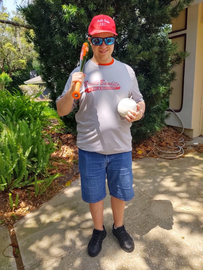 Greg Lindberg of Eyes Free Sports, holding a beep baseball and a baseball bat, wears a Braille Bandits jersey and hat to represent the Florida team he plays for.