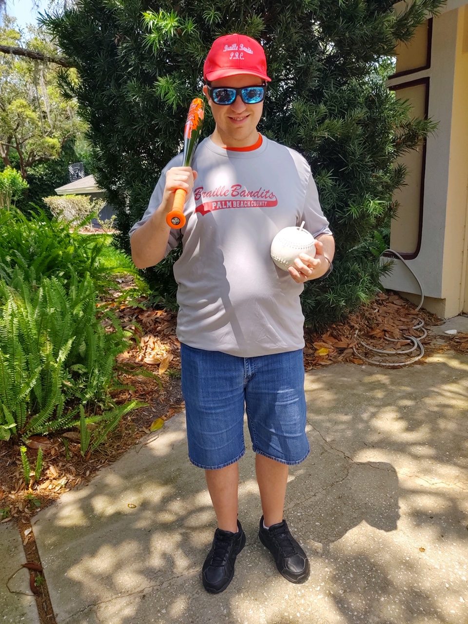 Greg holding a beep baseball and a baseball bat, wears a Braille Bandits jersey and hat to represent the Florida team he plays for.