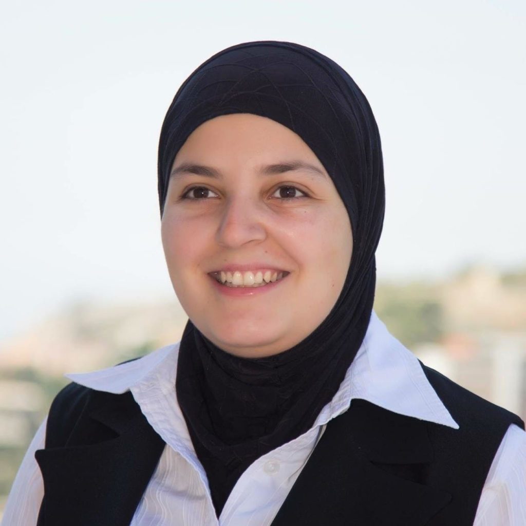 Mona Minkara, standing outdoors, wears a black hijab and white button-down-shirt and smiles broadly for the camera.