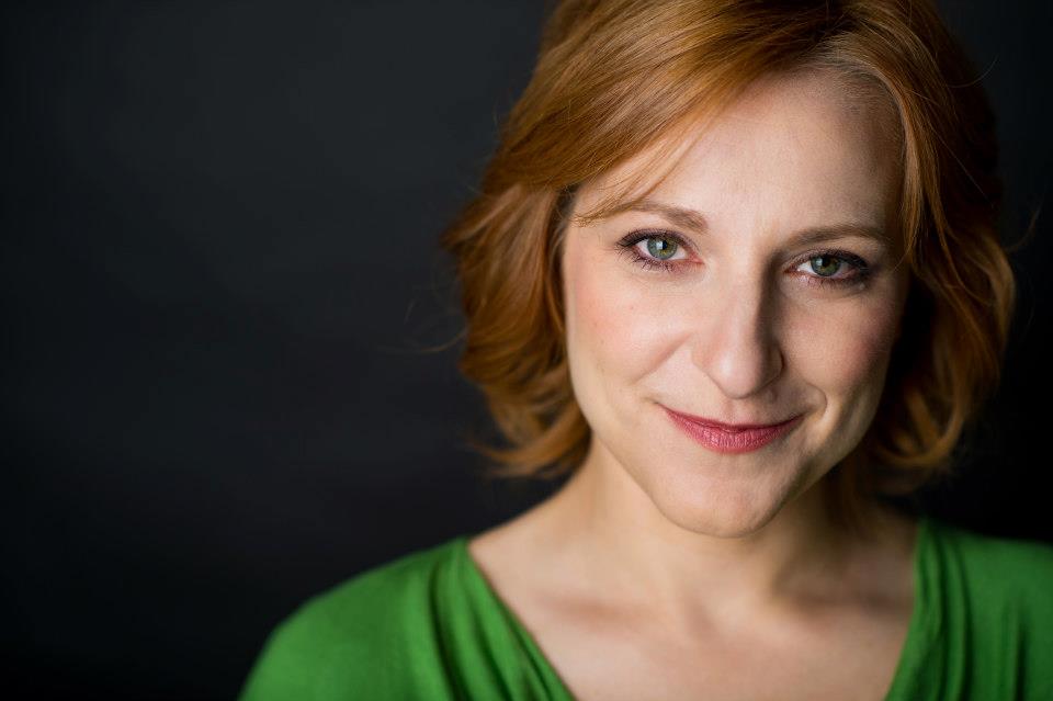 A professional headshot of Deana - she's standing against a black background wearing a green shirt and smiling.