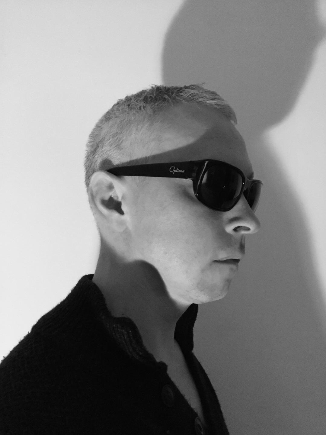 An artistic black-and-white photo of Clarke wearing a black shirt and black glasses. His shadow is cast at an upward angle on the white wall behind him.