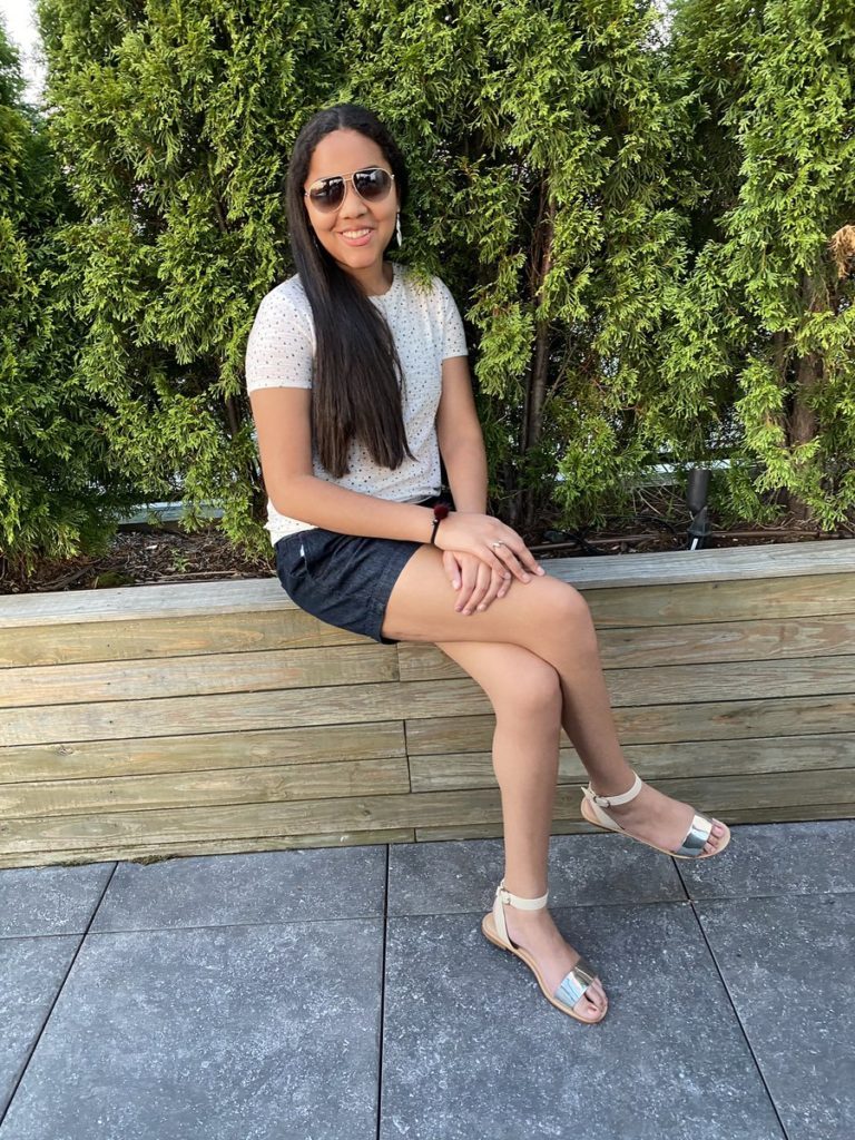 Gabby, smiling for the camera while wearing sunglasses and sitting on a low wall in front of greenery