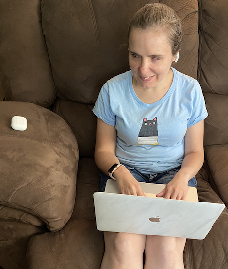 Kristy sits on a plush couch while working on a MacBook and smiling for the camera