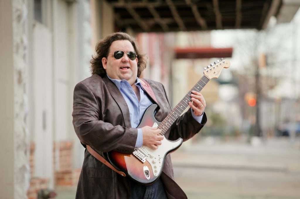Joey Stuckey stands on a sidewalk playing a Fender Stratocaster guitar