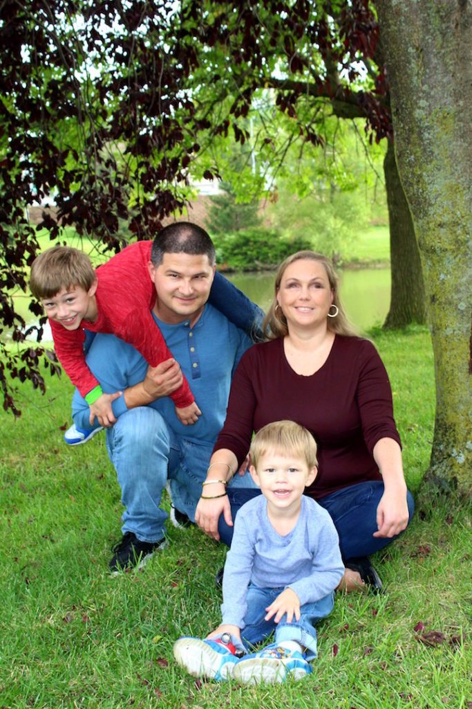 Bridgit and her family - husband Ross and sons Duncan and Declan - sit on green grass among trees, smiling for a family portrait