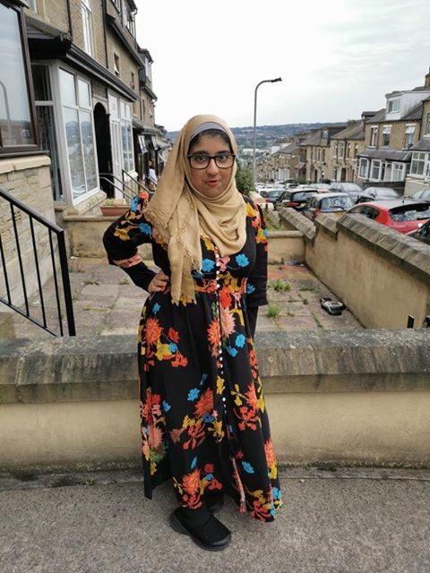 Ruqaiyah stands outdoors wearing black-framed glasses, a bright, floral dress and a head scarf. She has one hand on her hip and is looking confidently at the camera.