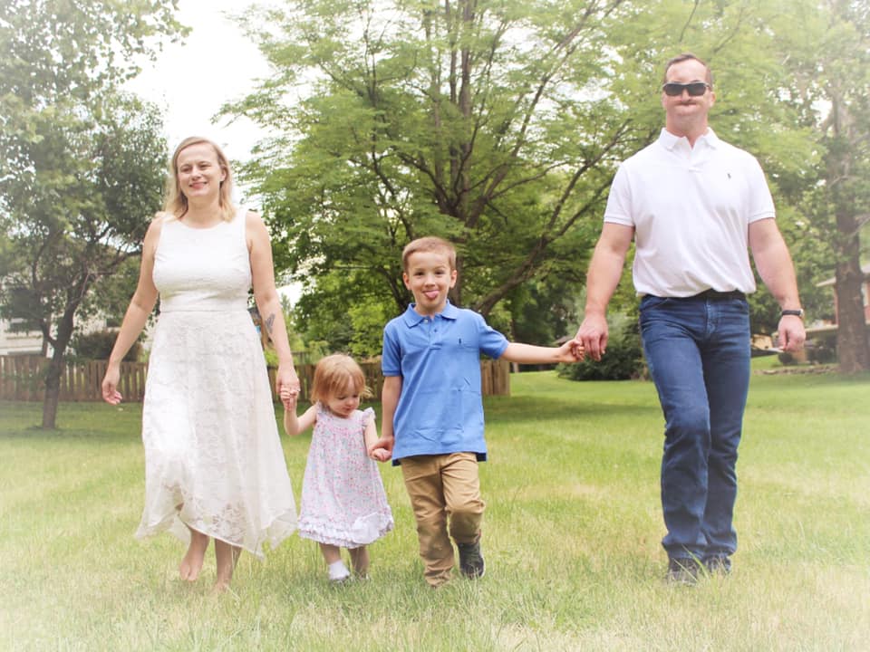  Stacy with her husband and two children, all holding hands and walking toward the camera, smiling, on a sunny day.