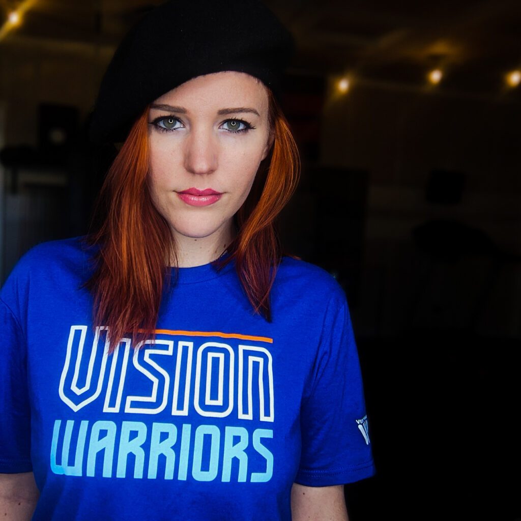 A young woman with mid-length reddish hair, wearing a black knit cap, bold pink lipstick, and a bright blue shirt that reads "Vision Warriors," looks confidently into the camera.