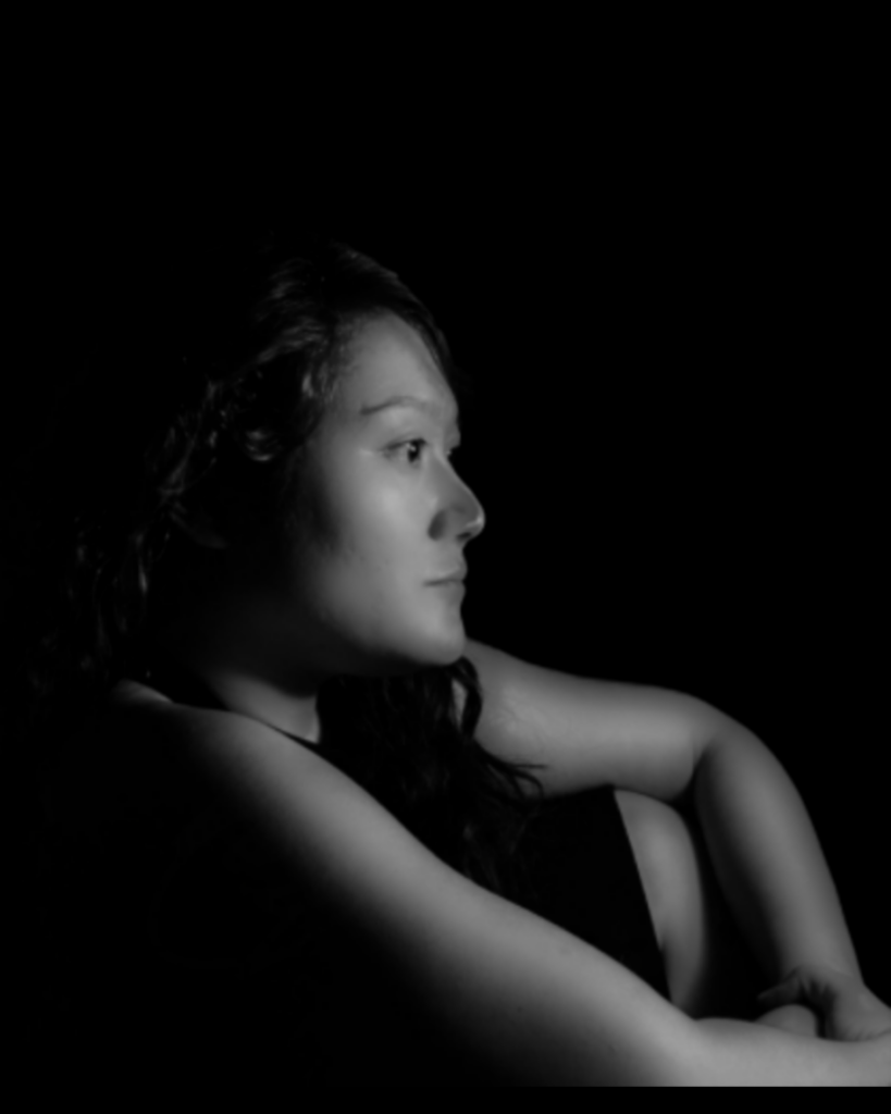 An artistic, black and white photo of Ana Cristina - against a black backdrop, she sits in profile, looking contemplative and determined