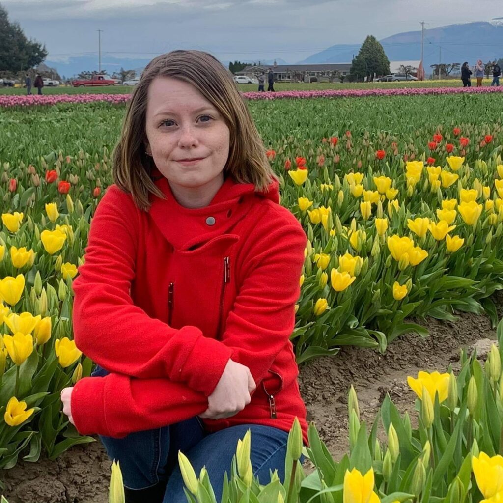 Courtney, a young woman with shoulder-length, light brown hair, kneels in a field of yellow and red flowers in bloom, smiling for the camera.