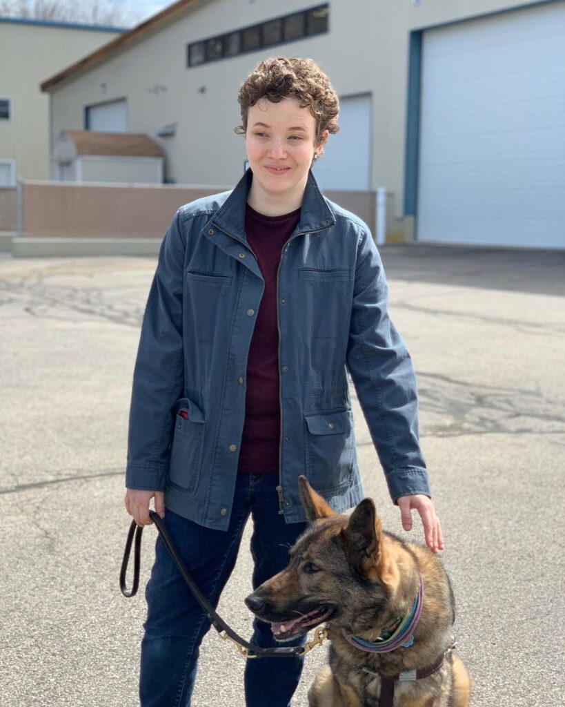 From Personal Growth to Pretzels: Lupin stands outdoors in an industrial parking lot, smiling for the camera. With his left hand, he's reaching down to pet Pluma, his German Shepherd guide dog.