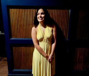 Precious Perez of RAMPD, a young latina with shoulder-length curly hair, smiles with her head tilted to the side in a dark room lit by a spotlight. She is wearing a bright yellow dress.