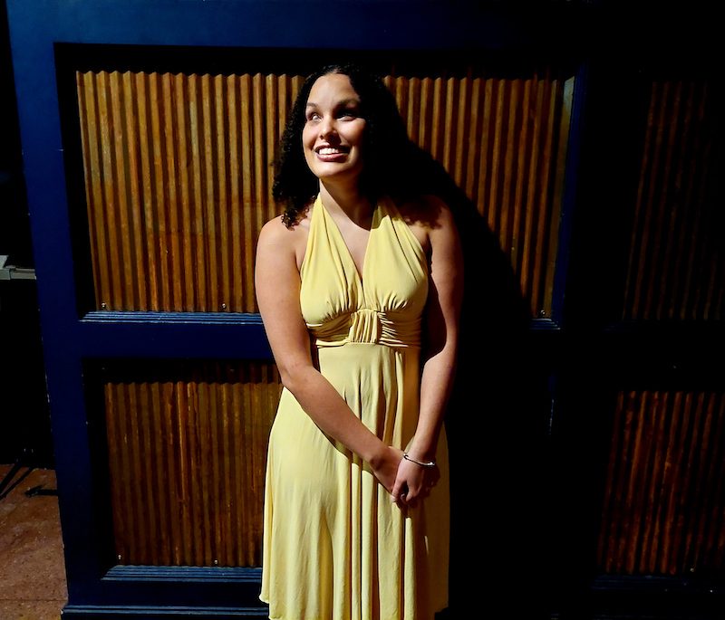Precious Perez of RAMPD, a young latina with shoulder-length curly hair, smiles with her head tilted to the side in a dark room lit by a spotlight. She is wearing a bright yellow dress.