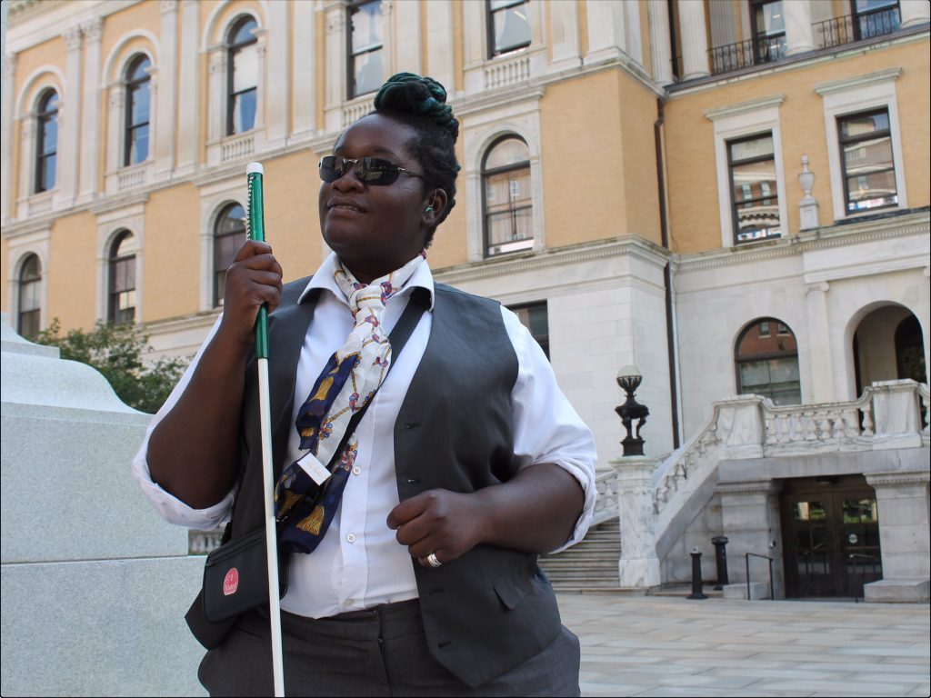 Casandra Xavier standing on a city street, holding her mobility cane and looking confident