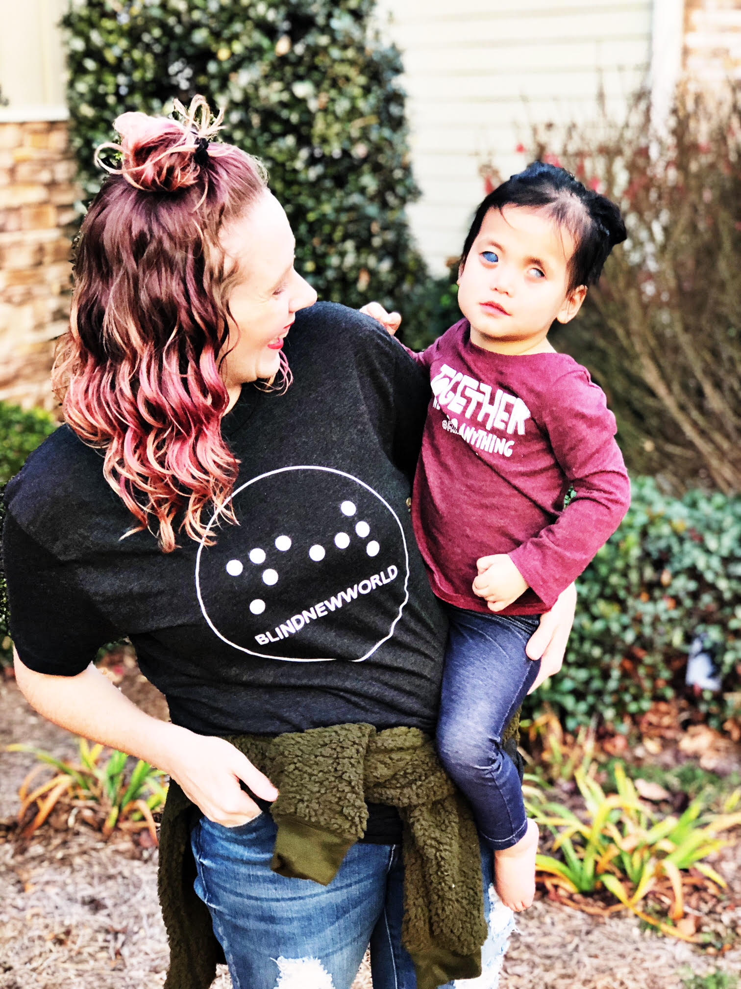 Eryn Austin, wearing a BlindNewWorld t-shirt, holds Primrose on her hip - facing her and smiling. Primrose is facing the camera and smiling.