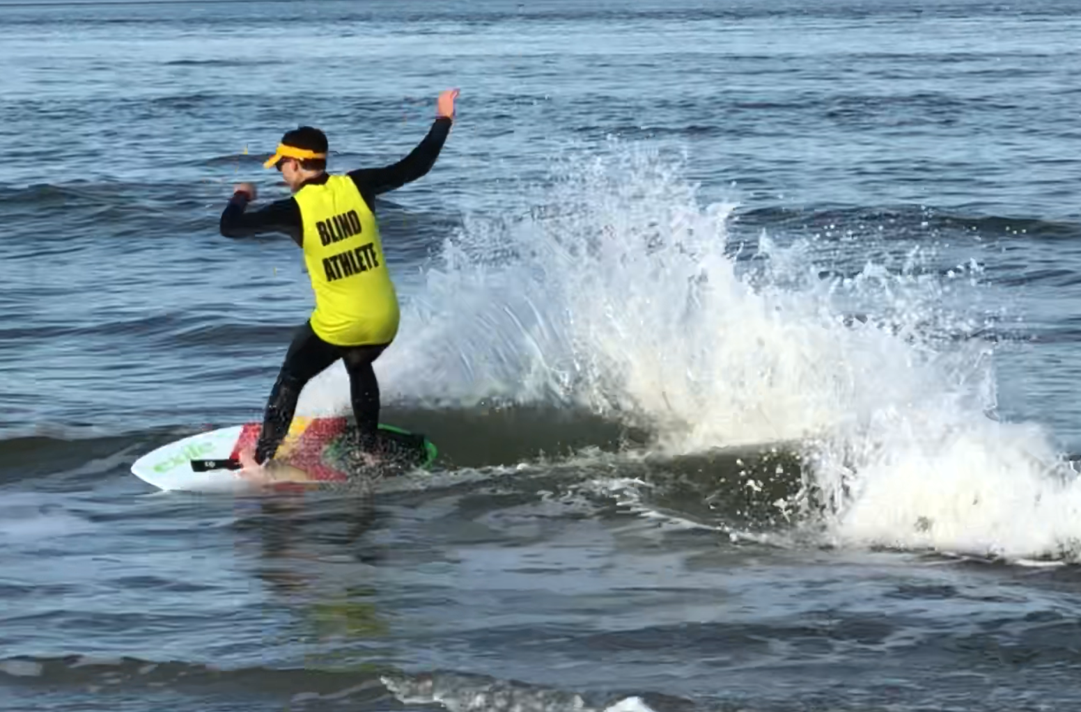 Kai Owens, skimboarding on an ocean wave. He's wearing black sunglasses, a bright yellow sun visor and a bright yellow jersey that reads BLIND ATHLETE over a black wetsuit.