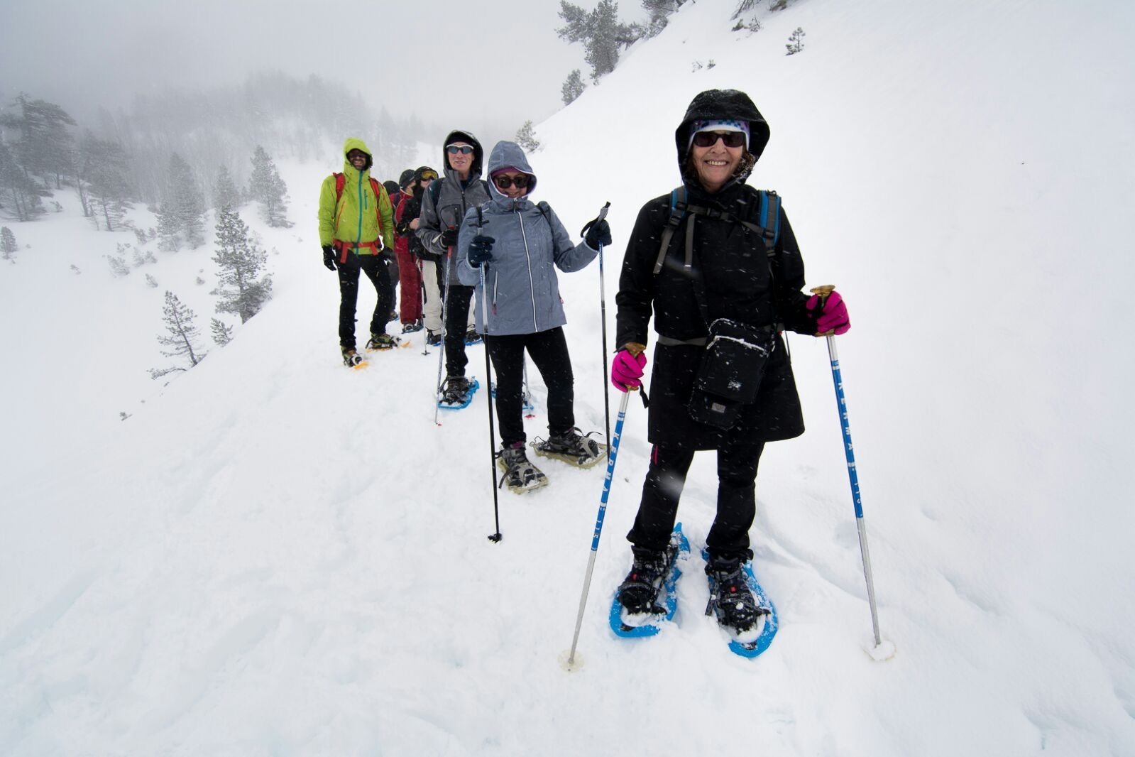 Wearing dark glasses, black snow gear, hot pink gloves and bright blue snowshoes, Begonia stands at the front of a group of snowshoers walking a trail on the side of a snowy mountain.