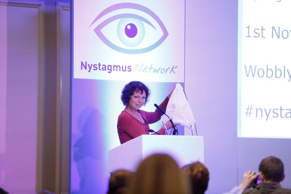 Sue Ricketts presenting on stage at a Nystagmus Network event. The stage is bathed in purple light and she stands at a podium in front of the Nystagmus Network logo.