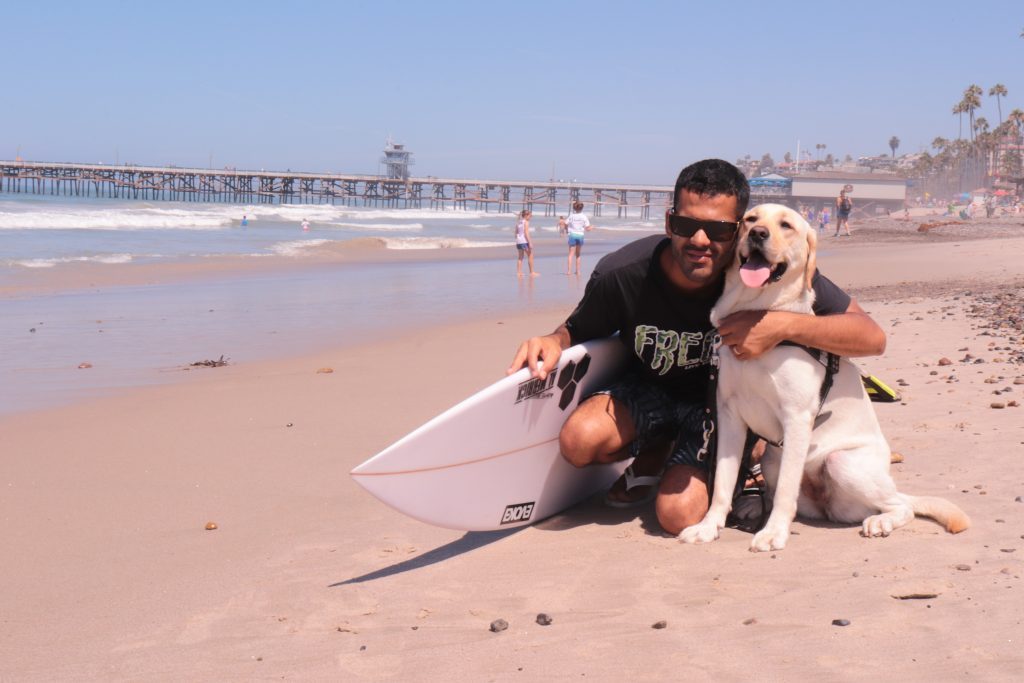 Derek Rabelo smiles while hugging his guide dog, Serenity, and holding a surfboard on a beach.