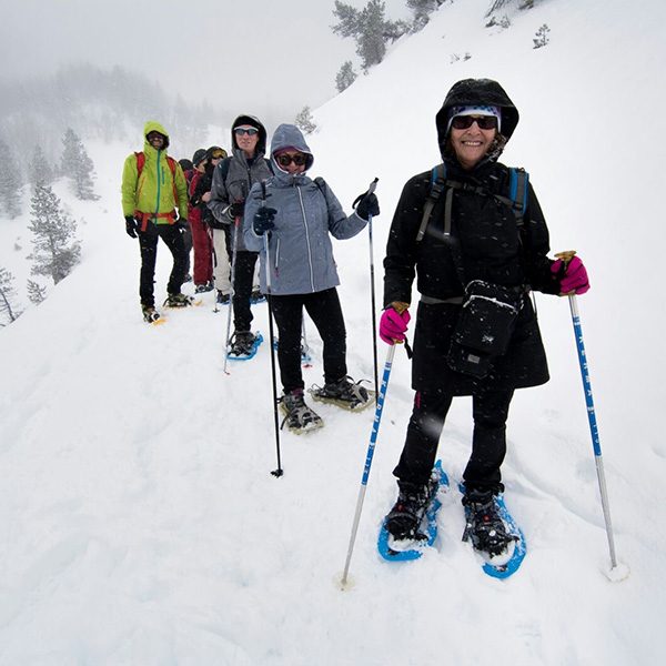 Wearing dark glasses, black snow gear, hot pink gloves and bright blue snowshoes, Begonia stands at the front of a group of snowshoers walking a trail on the side of a snowy mountain.