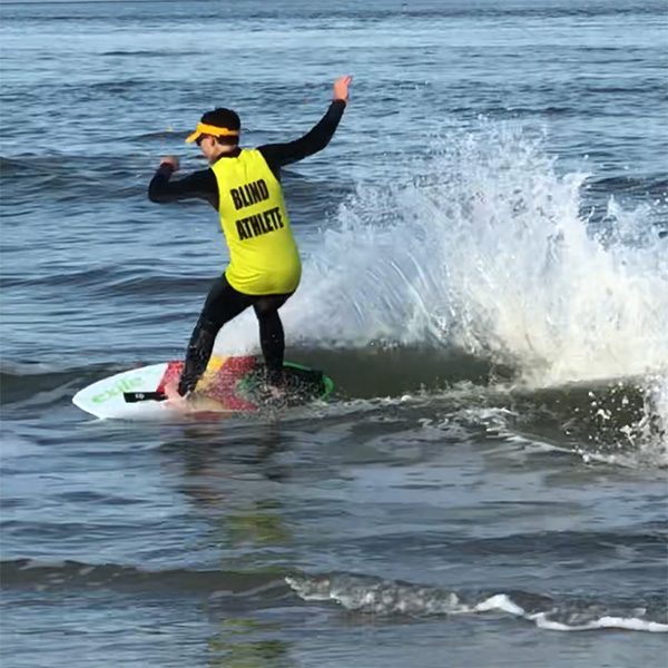 Kai Owens, skimboarding on an ocean wave. He's wearing black sunglasses, a bright yellow sun visor and a bright yellow jersey that reads BLIND ATHLETE over a black wetsuit.