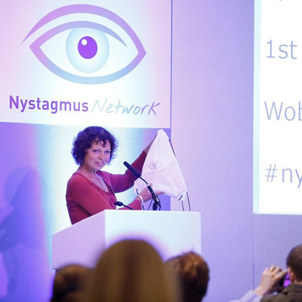 Sue Ricketts presenting on stage at a Nystagmus Network event. The stage is bathed in purple light and she stands at a podium in front of the Nystagmus Network logo.