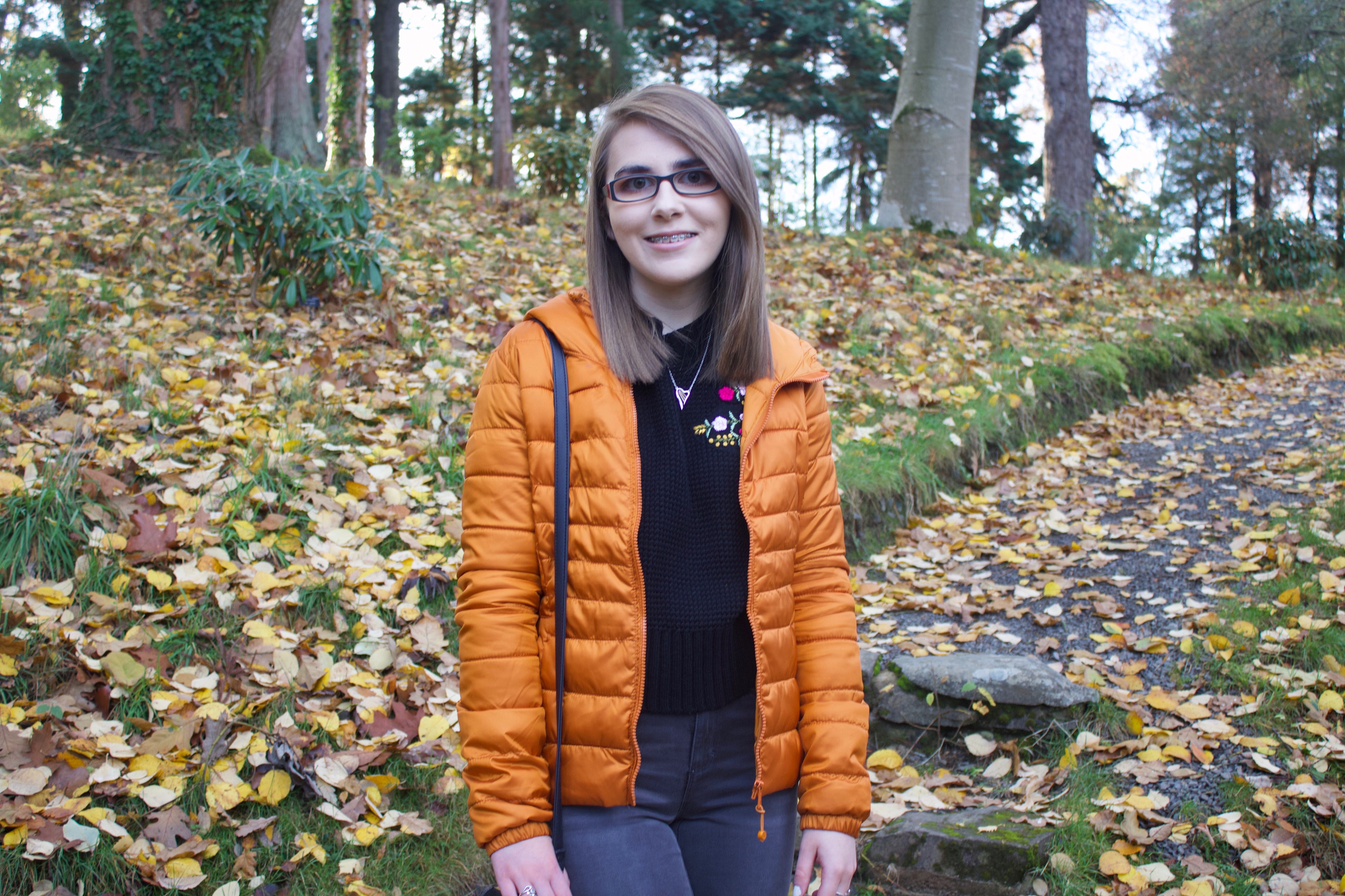 Elin stands smiling on a path in the woods. It's fall - she's surrounded by fallen leaves and she's wearing a warm, puffy orange coat.
