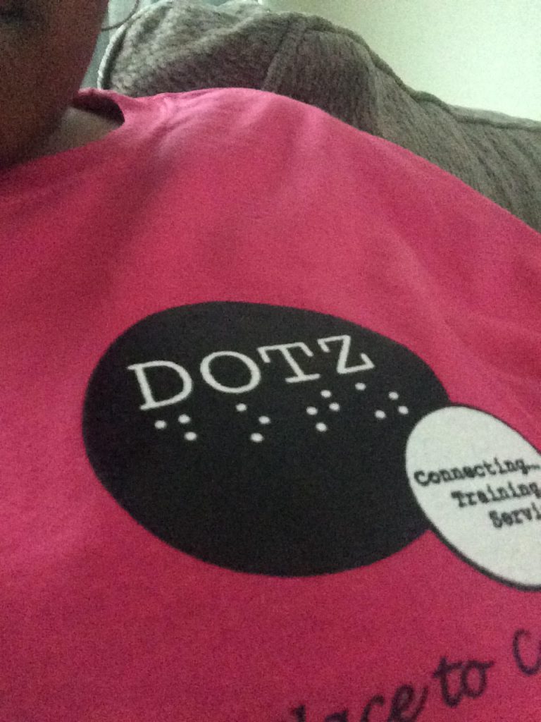 A close-up shot of Jasmyn's hot-pink t-shirt that reads "DOTZ" in text and braille.