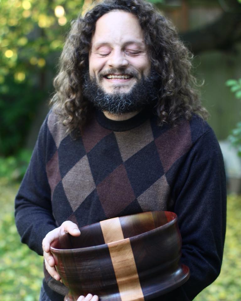 Man with dark beard and long dark curly hair (John) is wearing a dark sweater and holding a large wood bowl. The bowl is dark with a light colored stripe through the middle. Bowl is made of Black Walnut, Pedauk and Ash woods.