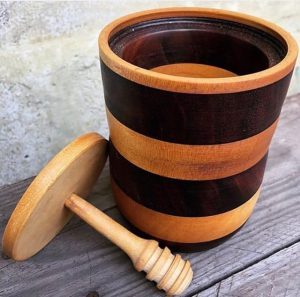 A striped wooden honey pot that has a lid next to it with a honey dipper attached.