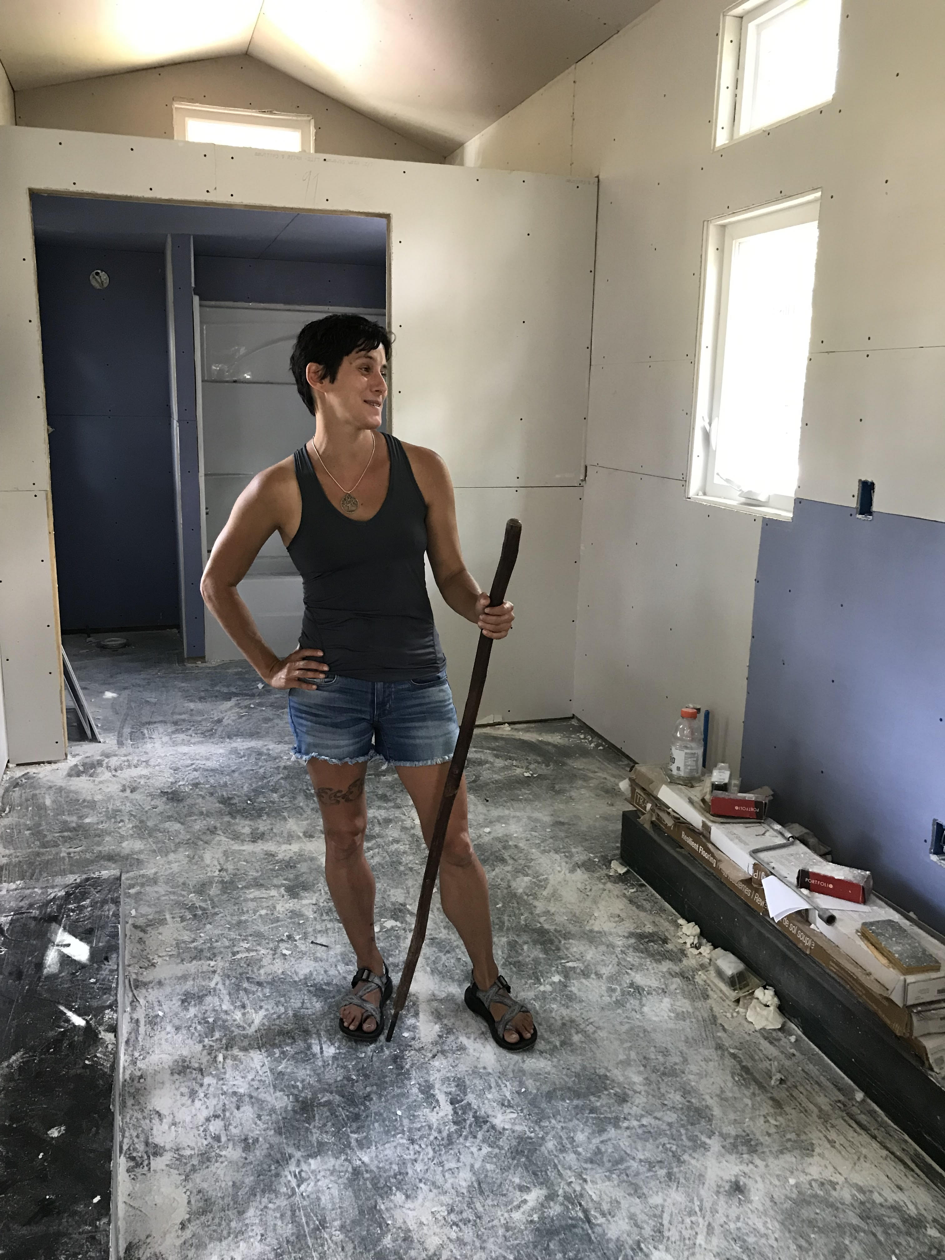 Gina Marie, holding a wooden walking stick, stands amid construction inside her tiny house.