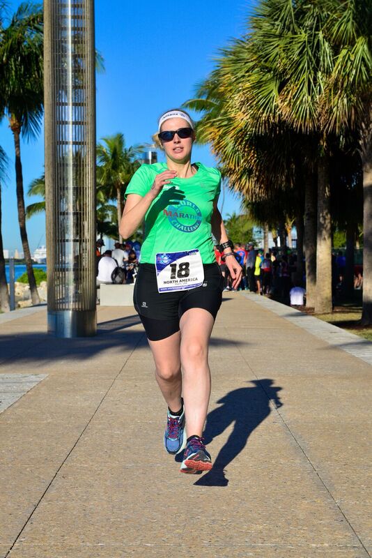 Sinead, wearing a bright green t-shirt, black shorts and dark glasses, runs confidently toward the camera. She's surrounded by palm trees. Photo credit: Mark Conlon, World Marathon Challenge