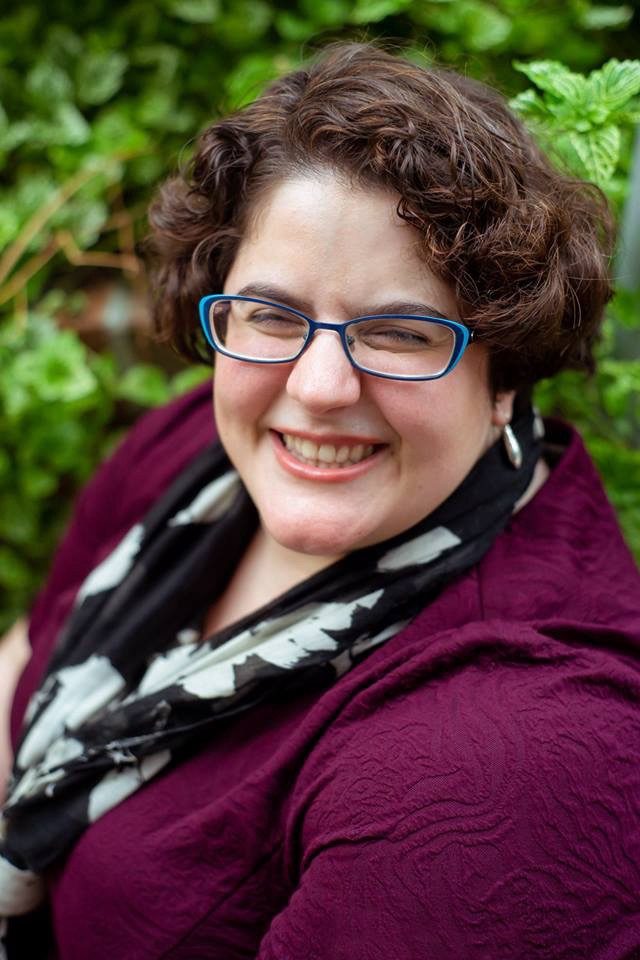Emily, posing for an outdoor professional headshot, wears a purple blazer, black-and-white floral scarf and blue-framed eyeglasses, and smiles joyfully for the camera. Photo credit: Chelsea Whitman