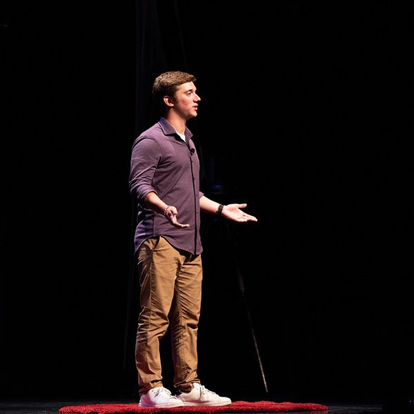  Louie, wearing a purple button-down and khakis, stands in his “red dot” on the TEDx stage
