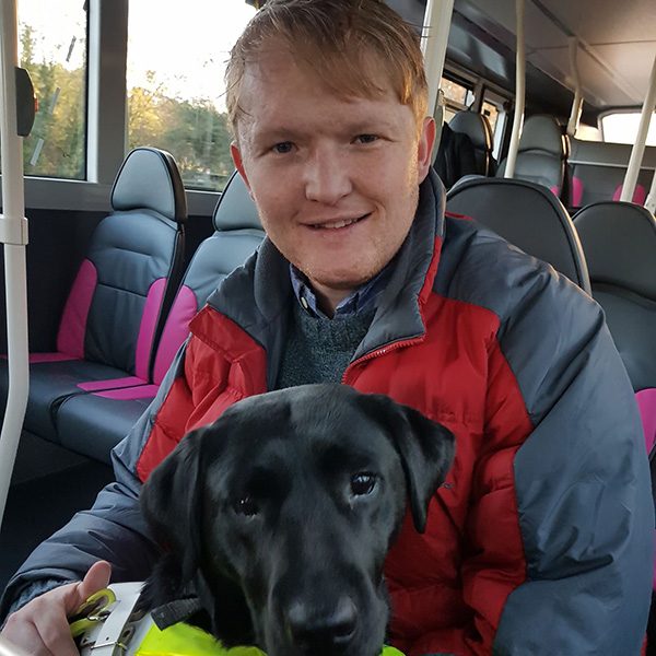 Nathan smiles for the camera while riding the bus. His guide dog, a black lab named Maisie, sits in his lap and looks at the camera.