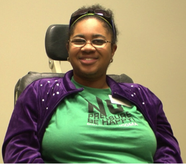 Jasmyn, wearing a green t-shirt and purple cardigan, smiles broadly for the camera.
