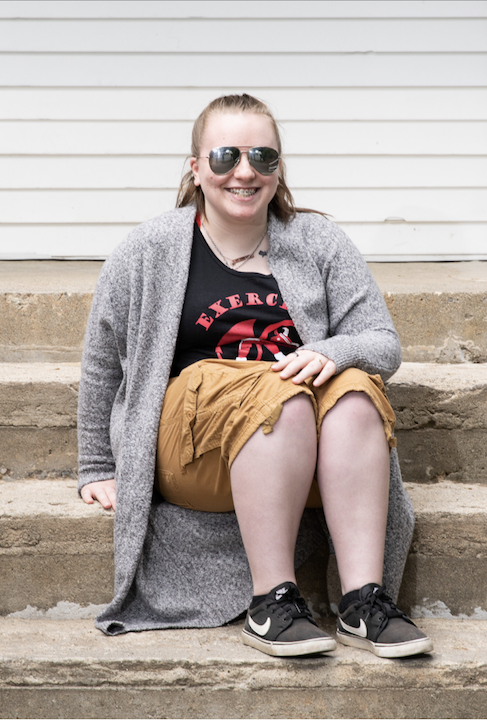 Ashley sits outdoors on concrete steps. She's wearing sunglasses and smiling for the camera.