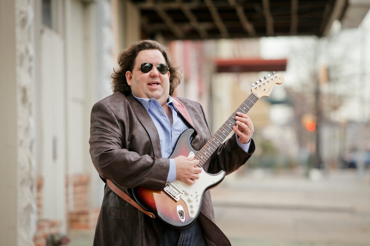 Joey Stuckey stands on a sidewalk playing a Fender Stratocaster guitar